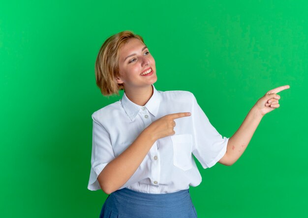 Молодая улыбающаяся русская блондинка указывает в сторону, изолированную на зеленом фоне с копией пространства