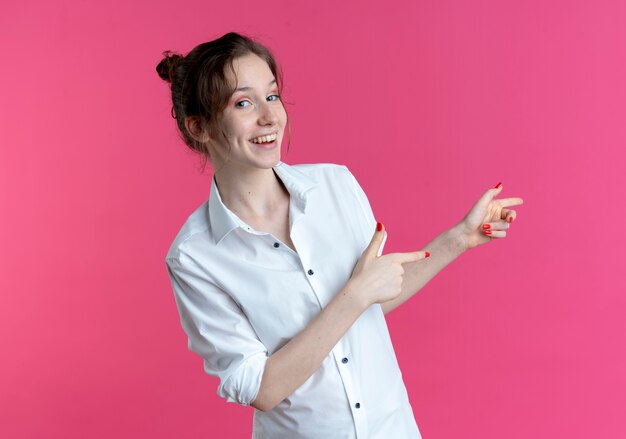 복사 공간 핑크 공간에 고립 된 두 손으로 측면에서 젊은 웃는 금발 러시아 여자 포인트