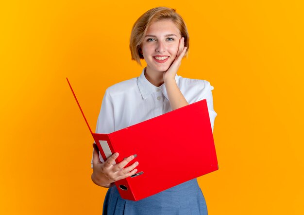 Young smiling blonde russian girl puts hand on face holding file folder isolated on orange background with copy space