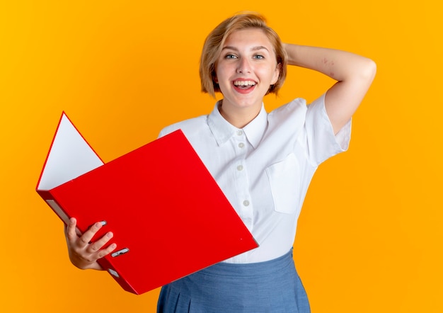young smiling blonde russian girl puts hand on head behind holding file folder isolated on orange background with copy space