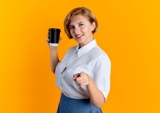 young smiling blonde russian girl stands sideways holding coffee cup and pointing at camera