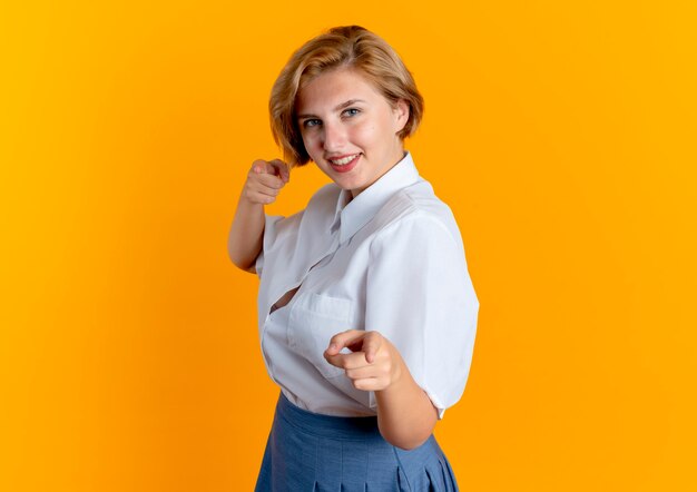 Young smiling blonde russian girl stands sideways pointing at camera isolated on orange background with copy space