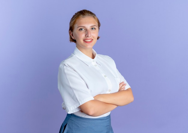 Young smiling blonde russian girl stands with crossed arms isolated on purple background with copy space