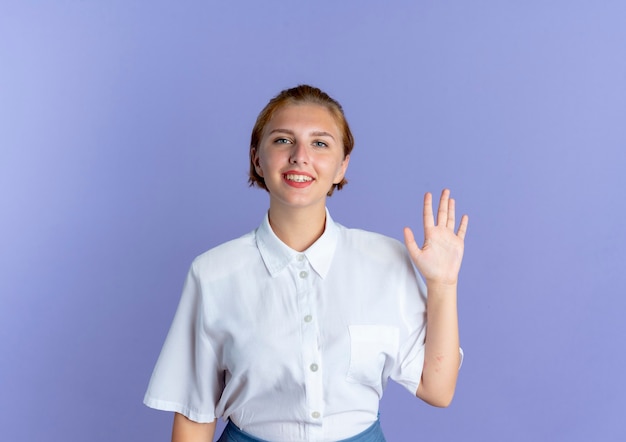 Young smiling blonde russian girl stands with raised hand isolated on purple background with copy space