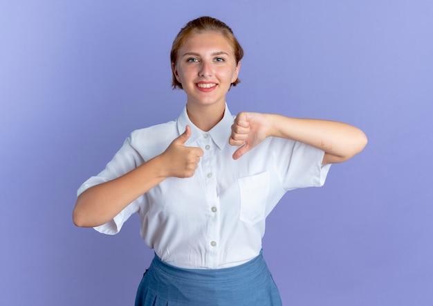 Young smiling blonde russian girl thumbs up and thumbs down isolated on purple background with copy space