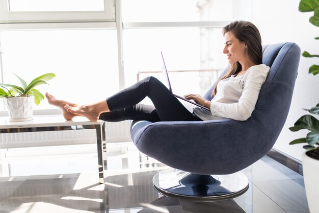젊은 미소 갈색 머리 소녀 편안한 분위기에서 노트북에서 일하는 집에서 가벼운 아늑한 방에 창 근처에 현대 의자에 앉아있다
