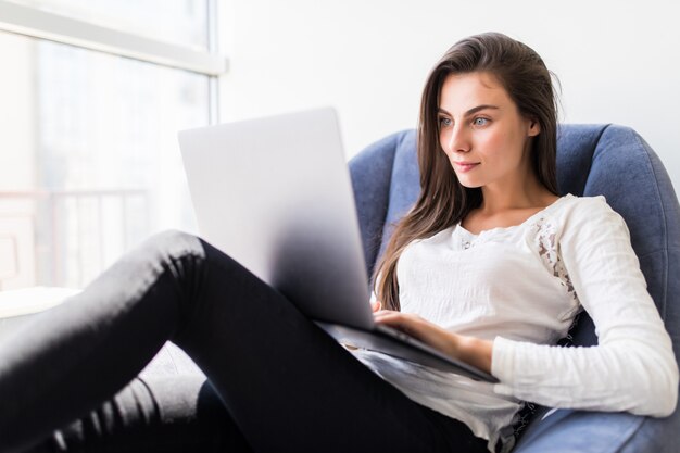 젊은 미소 갈색 머리 소녀 편안한 분위기에서 노트북에서 일하는 집에서 가벼운 아늑한 방에 창 근처에 현대 의자에 앉아있다