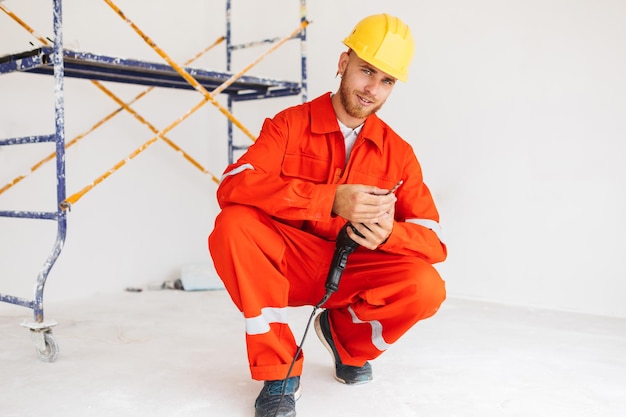 Giovane costruttore sorridente in abiti da lavoro arancioni e elmetto protettivo giallo che guarda sognante nella fotocamera con in mano un trapano elettrico con impalcature sullo sfondo