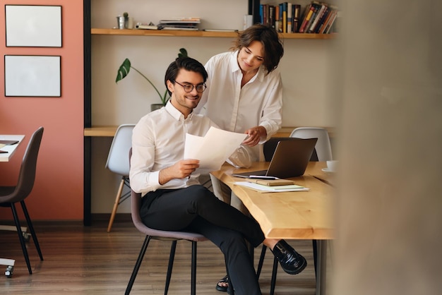Молодые улыбающиеся коллеги по бизнесу с удовольствием работают с бумагами в современном офисе