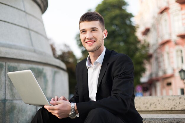 Молодой улыбающийся бизнесмен в классическом черном костюме и белой рубашке с беспроводными наушниками счастливо смотрит в сторону, держа ноутбук в руках, проводя время на свежем воздухе