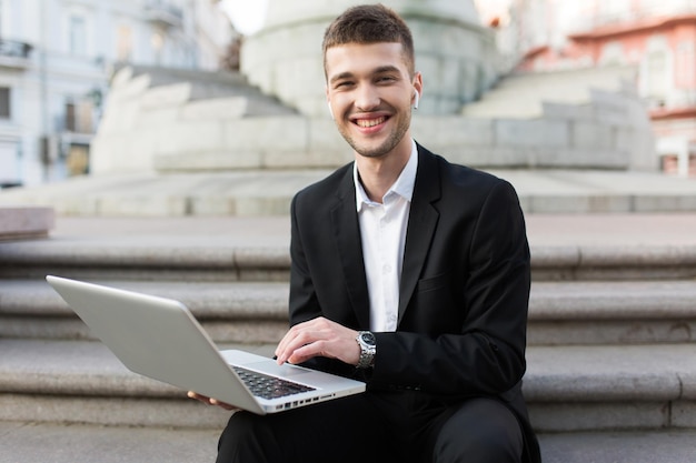 Молодой улыбающийся бизнесмен в классическом черном костюме с беспроводными наушниками радостно смотрит в камеру, держа ноутбук в руках, проводя время на свежем воздухе