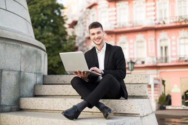 Young smiling businessman in classic black suit with wireless earphones joyfully looking in camera working with laptop on little stairs while spending time outdoor