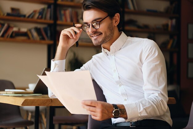 안경과 흰색 셔츠를 입은 웃고 있는 젊은 사업가가 현대 사무실에서 서류로 행복하게 일하고 있다