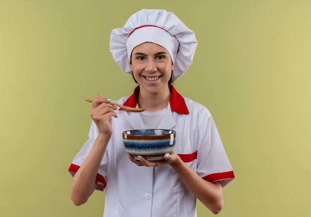 Молодая улыбающаяся кавказская девушка-повар в униформе шеф-повара держит миску и ложку на зеленом с копией пространства