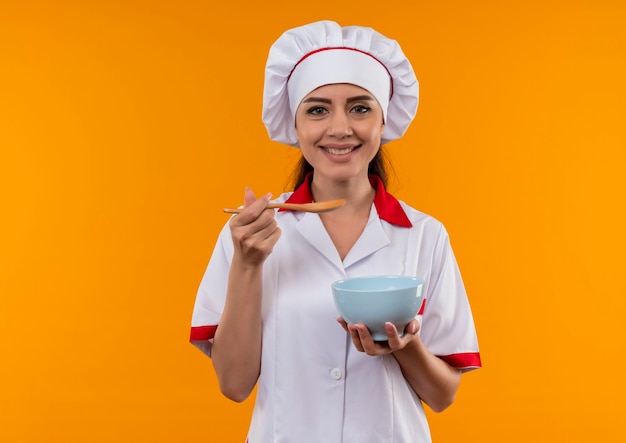 요리사 유니폼에 젊은 웃는 백인 요리사 소녀 보유 그릇과 오렌지 벽 복사 공간에 고립 된 나무 숟가락