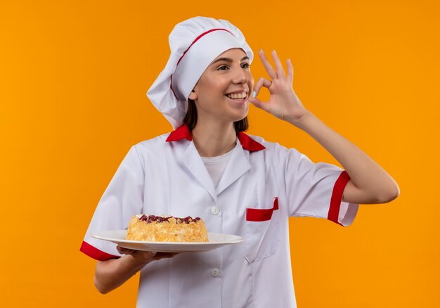 シェフの制服を着た若い笑顔の白人料理人の女の子は、プレートにケーキを保持し、コピースペースとオレンジ色のスペースで隔離のおいしいサインをジェスチャー