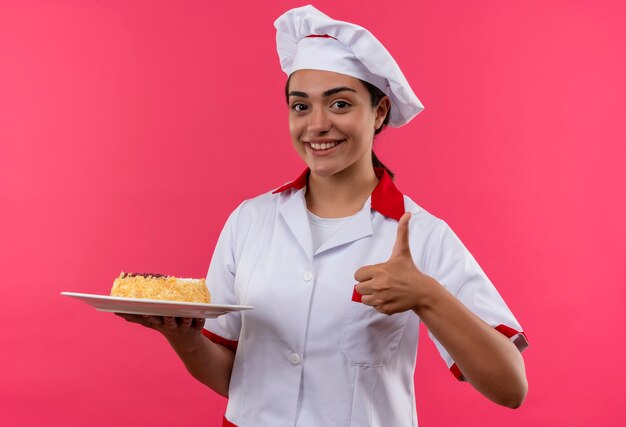 Young smiling caucasian cook girl in chef uniform holds cake on plate and thumbs up isolated on pink wall with copy space