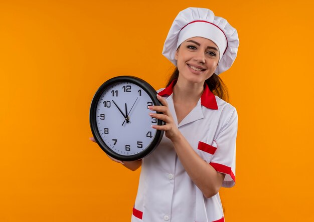 요리사 유니폼에 젊은 웃는 백인 요리사 소녀 보유 시계 복사 공간이 오렌지 벽에 고립