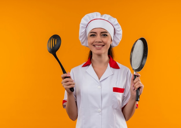 Young smiling caucasian cook girl in chef uniform holds frying pan and spatula isolated on orange wall with copy space