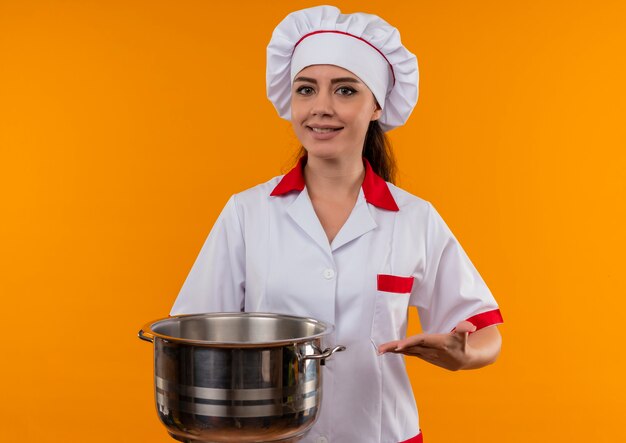 Young smiling caucasian cook girl in chef uniform holds and points with hand at pot isolated on orange wall with copy space