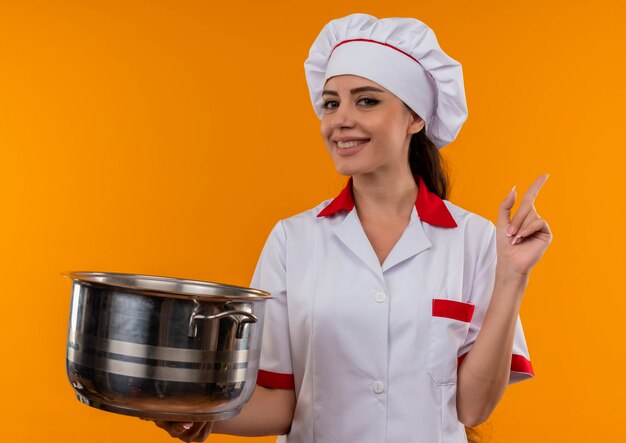 Young smiling caucasian cook girl in chef uniform holds pot and points up isolated on orange wall with copy space