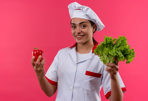 Young smiling caucasian cook girl in chef uniform holds red pepper and salad isolated on pink wall with copy space