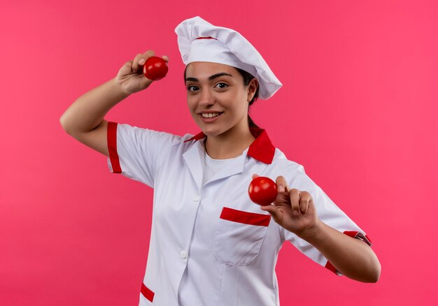 요리사 유니폼에 젊은 웃는 백인 요리사 소녀 복사 공간이 분홍색 벽에 고립 된 토마토를 보유