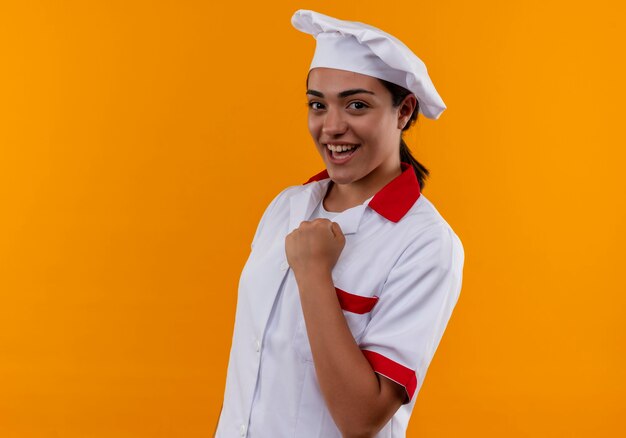 シェフの制服を着た若い笑顔の白人料理人の女の子は、オレンジ色の壁に拳を隔離し続けます