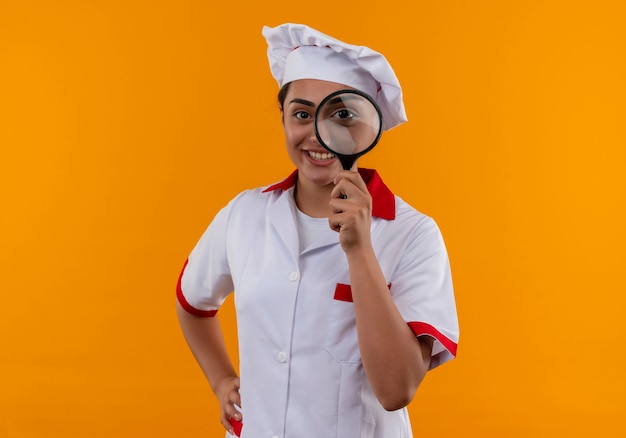 Young smiling caucasian cook girl in chef uniform looks through magnifying glass or loupe isolated on orange wall 