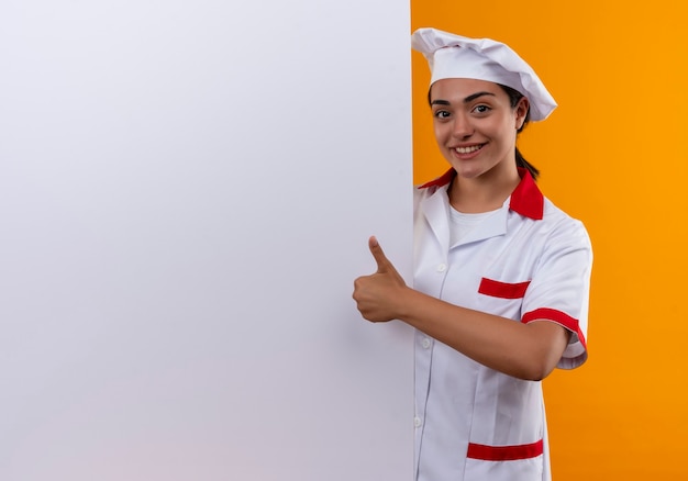 Молодая улыбающаяся кавказская девушка-повар в униформе шеф-повара стоит за белой стеной и показывает палец вверх, изолированную на оранжевой стене с копией пространства
