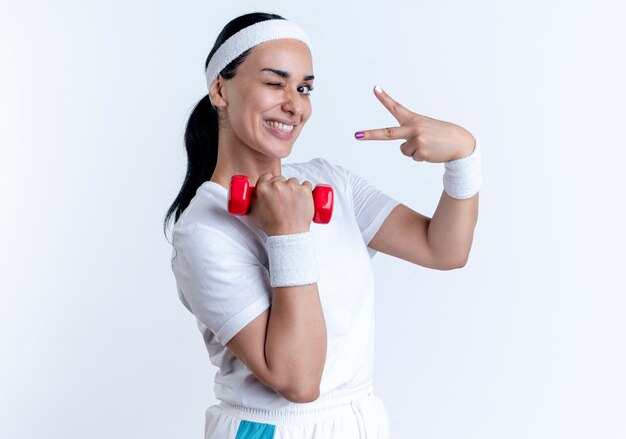 Young smiling caucasian sporty woman wearing headband and wristbands blinks eye holding dumbbell and gesturing victory hand sign isolated on white space with copy space