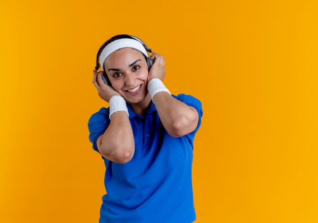 Young smiling caucasian sporty woman wearing headband and wristbands holds headphones on orange  with copy space