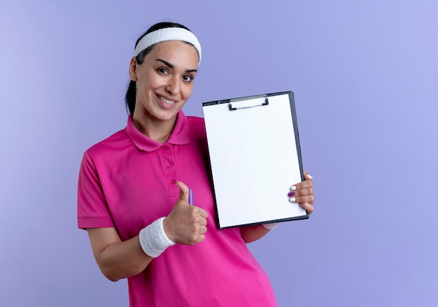 Молодая улыбающаяся кавказская спортивная женщина с головной повязкой и браслетами показывает палец вверх, держа ручку и буфер обмена, изолированные на фиолетовом фоне с копией пространства