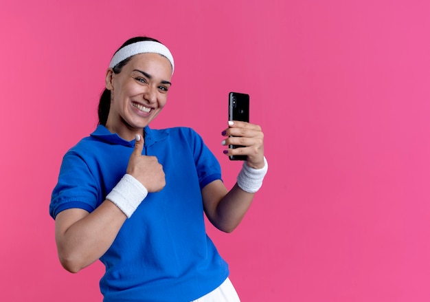 Молодая улыбающаяся кавказская спортивная женщина в головной повязке и браслетах показывает палец вверх, держа телефон на розовом фоне с копией пространства