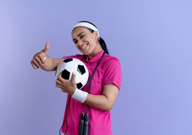 Young smiling caucasian sporty woman wearing headband and wristbands with jumping rope on neck holds ball thumbs up on purple  with copy space
