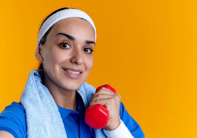 Young smiling caucasian sporty woman wearing headband and wristbands with towel on neck holds dumbbell on orange  with copy space