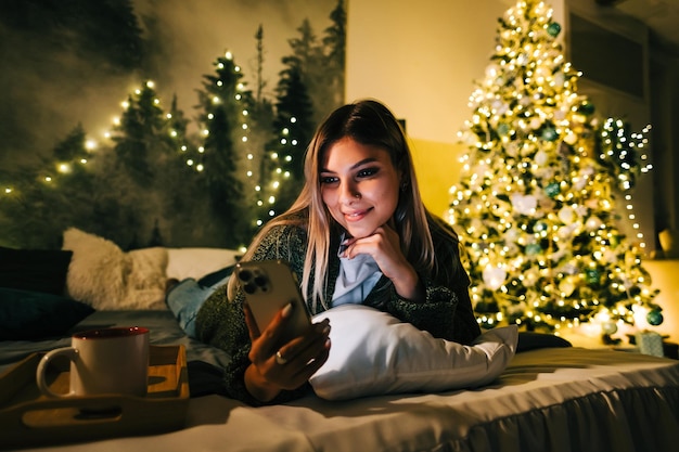 휴일에 집에서 침대에서 휴대전화를 사용하여 웃고 있는 젊은 백인 여성.