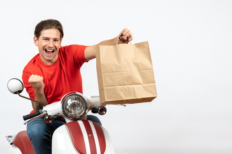 Free Photo young smiling confident courier guy in red uniform sitting on scooter holding paper bag on white wall