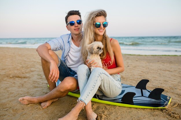 Young smiling couple having fun on beach sitting on sand with surf boards playing with dog