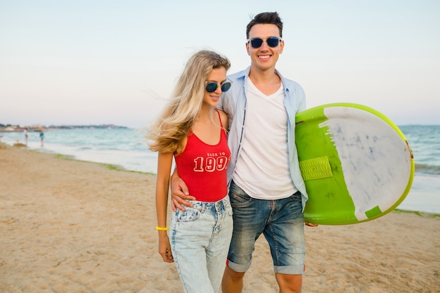 Young smiling couple having fun on beach walking with surf board