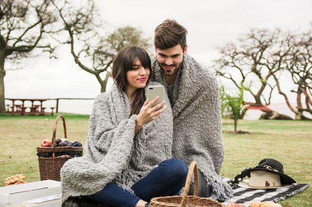 Молодая пара улыбаясь, глядя на мобильный телефон в парке