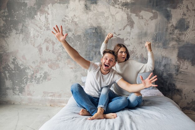 Молодая улыбающаяся пара, сидящая на кровати дома в повседневной одежде, мужчина и женщина, веселятся вместе, сумасшедшие положительные эмоции, счастливы, держась за руки