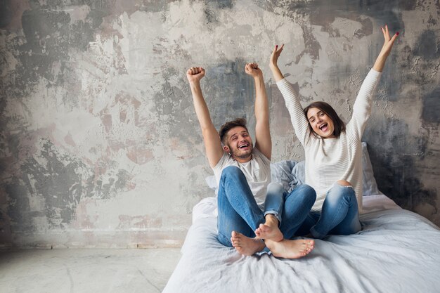 Young smiling couple sitting on bed at home in casual outfit, man and woman having fun together, crazy positive emotion, happy, holding hands up