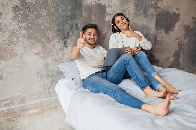 Молодая улыбающаяся пара, сидящая на кровати дома в повседневной одежде, человек, работающий внештатно на ноутбуке, женщина, слушающая музыку в наушниках, проводя счастливое время вместе, положительные эмоции, глядя в камеру