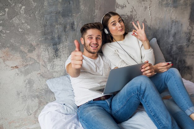 Young smiling couple sitting on bed at home in casual outfit, man working freelance on laptop, woman listening to music on headphones, spending happy time together, positive emotion, looking in camera
