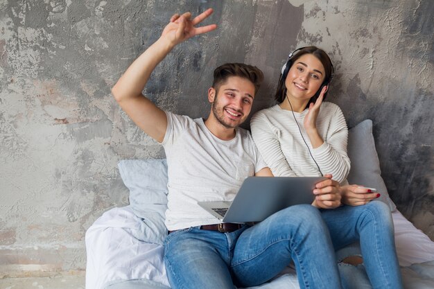 Young smiling couple sitting on bed at home in casual outfit, man working freelance on laptop, woman listening to music on headphones, spending happy time together, positive emotion, looking in camera