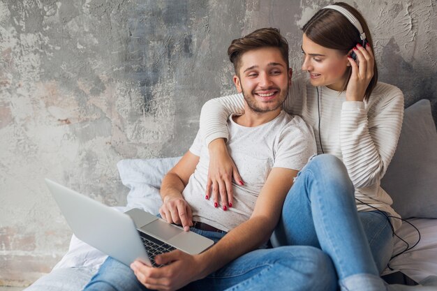 Young smiling couple sitting on bed at home in casual outfit, man working freelance on laptop, woman listening to music on headphones, spending happy time together