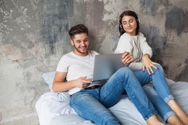 Young smiling couple sitting on bed at home in casual outfit, man working freelance on laptop, woman listening to music on headphones, spending time together