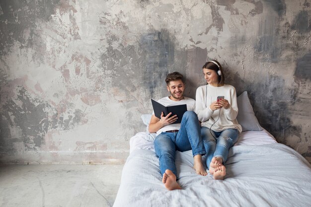 Young smiling couple sitting on bed at home in casual outfit reading book wearing jeans, man reading book, woman listening to music on headphones, spending romantic time together