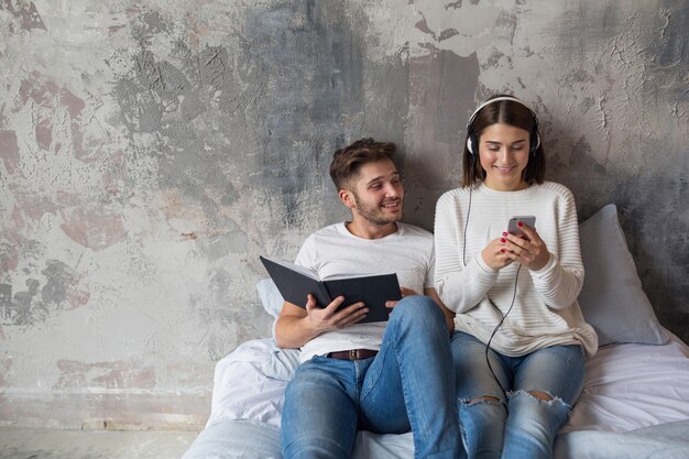 Young smiling couple sitting on bed at home in casual outfit reading book wearing jeans, man reading book, woman listening to music on headphones, spending romantic time together